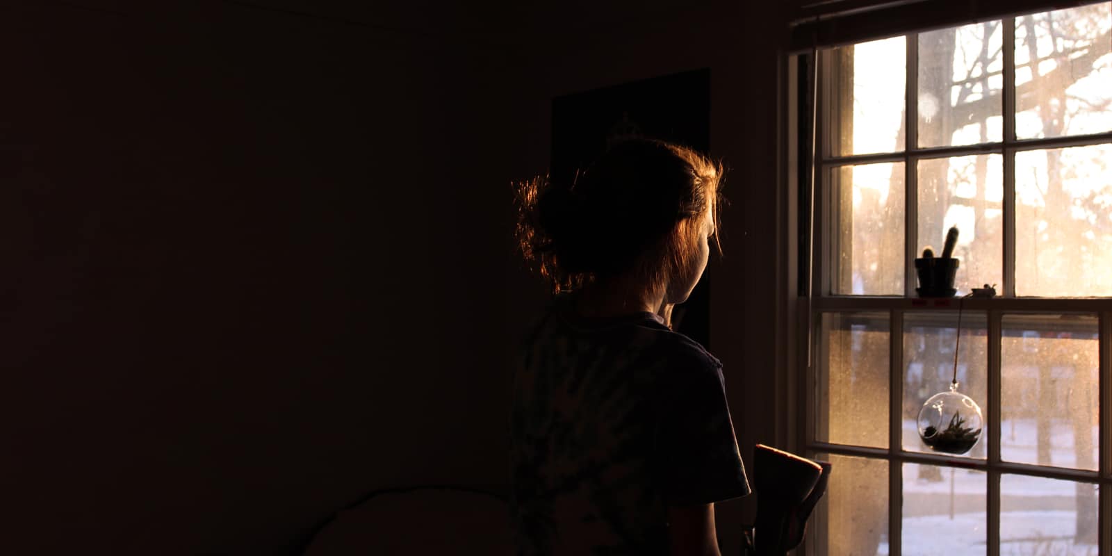 An image of a woman looking out the window in a darkly lit room