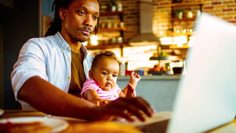 a young father sits at his laptop with his young child in his lap