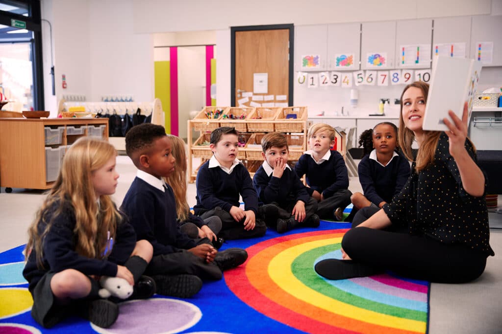 Teaching Assistant with pupils sitting on a rainbow rug.