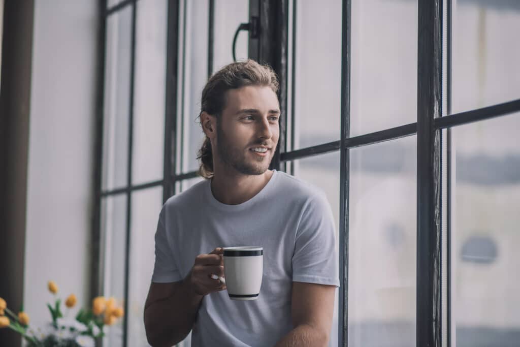 A man smiling and looking out the window in the morning, with a coffee in his hand.