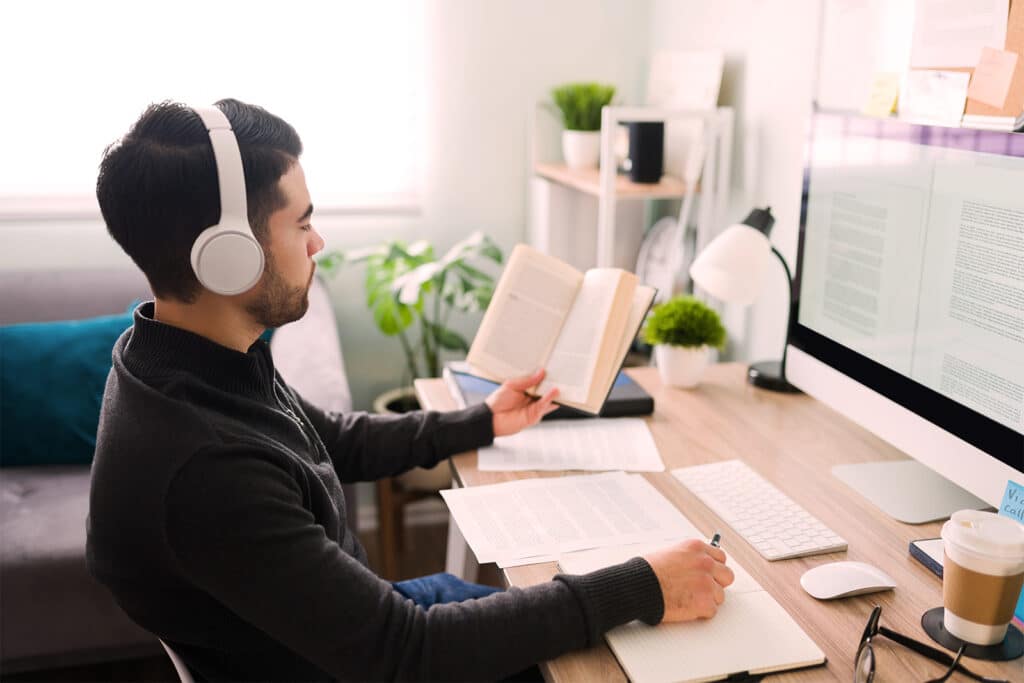 Man sat in front of a Mac with headphones on, reading a book and writing.