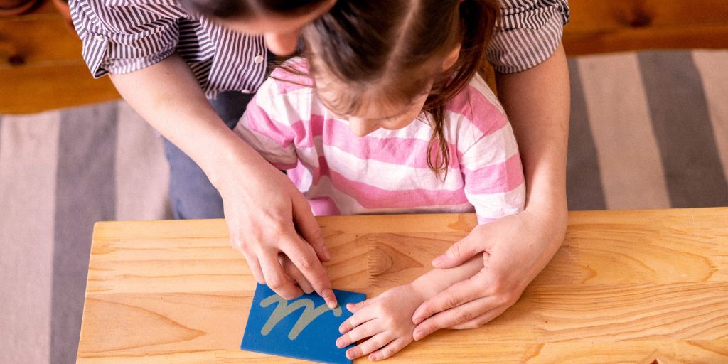 A mother teacher her daughter sensory play by pushing her finger along a sheet of card with a sandpaper embossed letter M