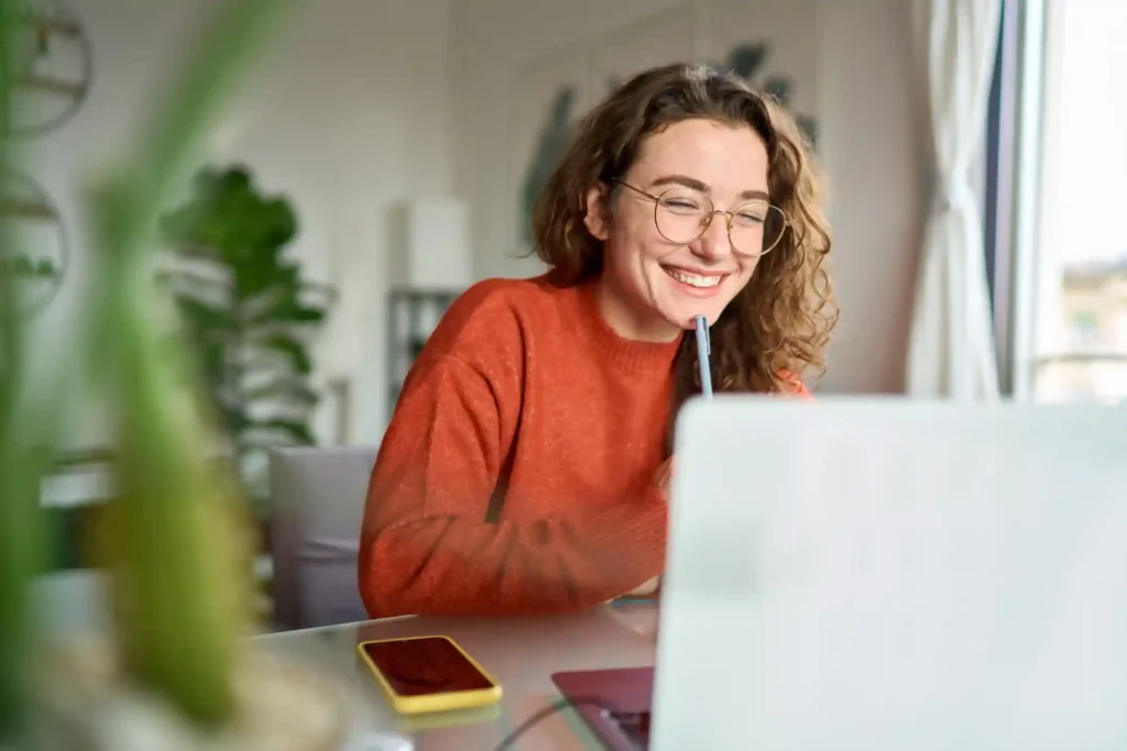 Happy young woman using laptop sitting at desk writing notes while watching webinar.